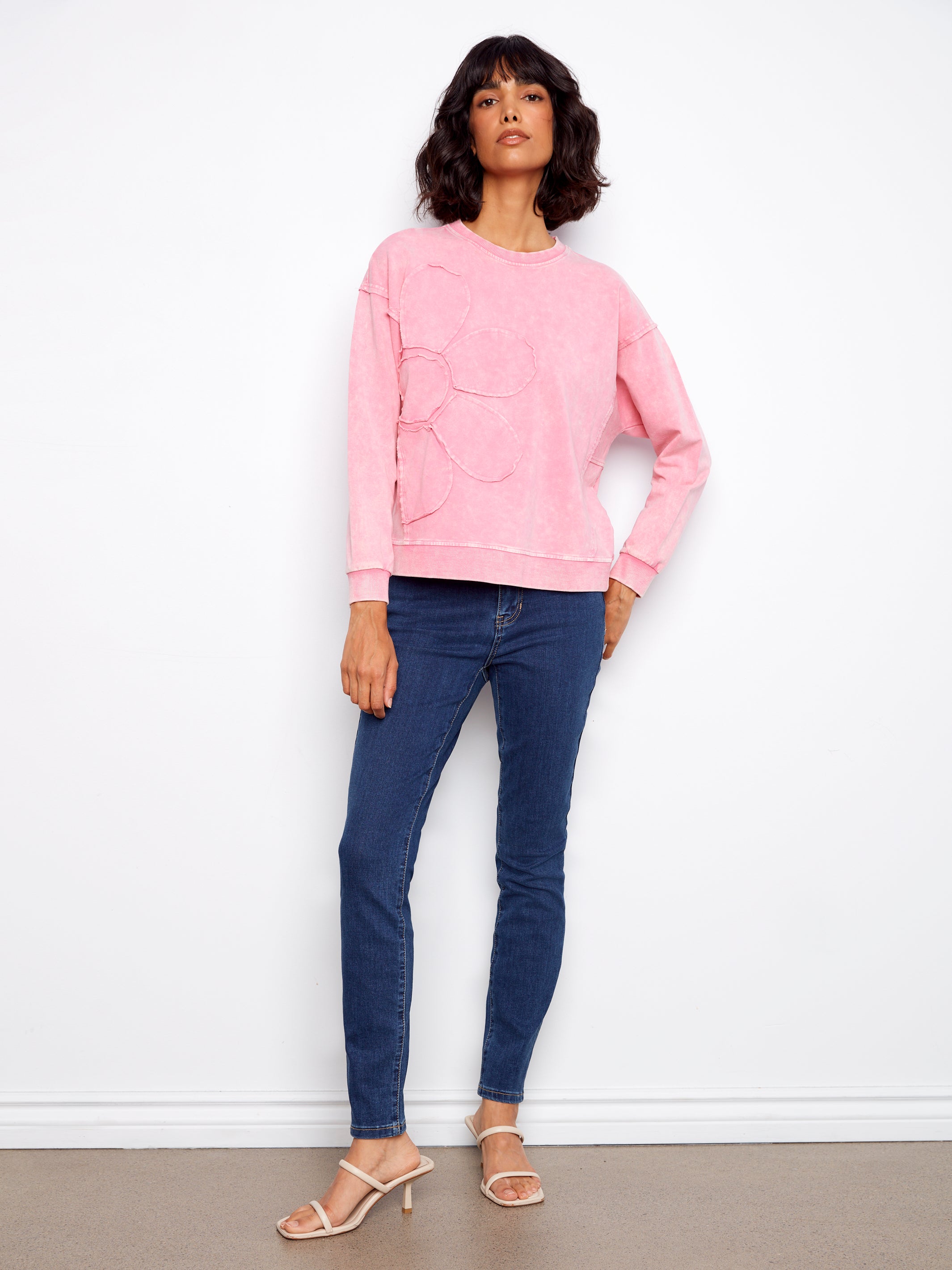 Woman wearing a pink sweatshirt and blue jeans against a white wall