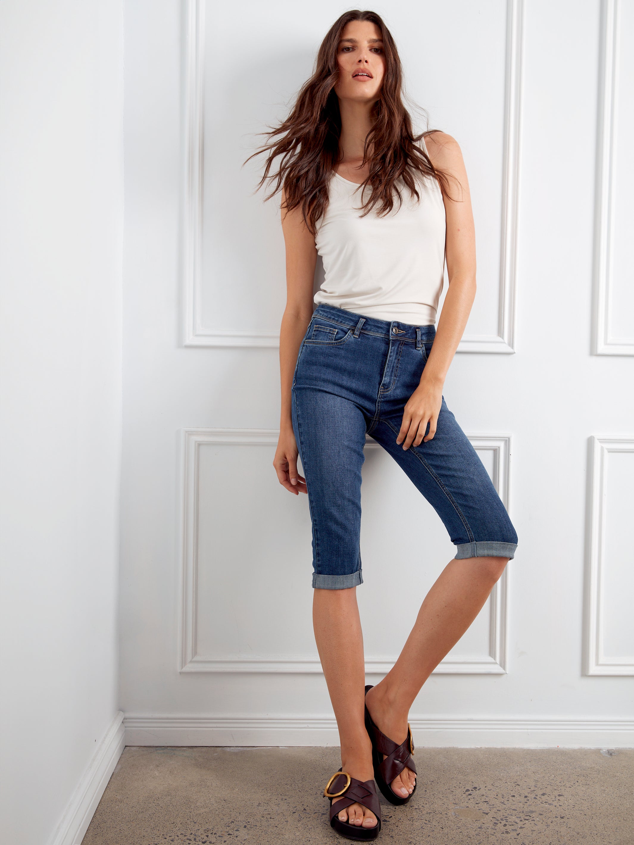 Woman wearing a white tank top and blue jeans standing against a white paneled wall.