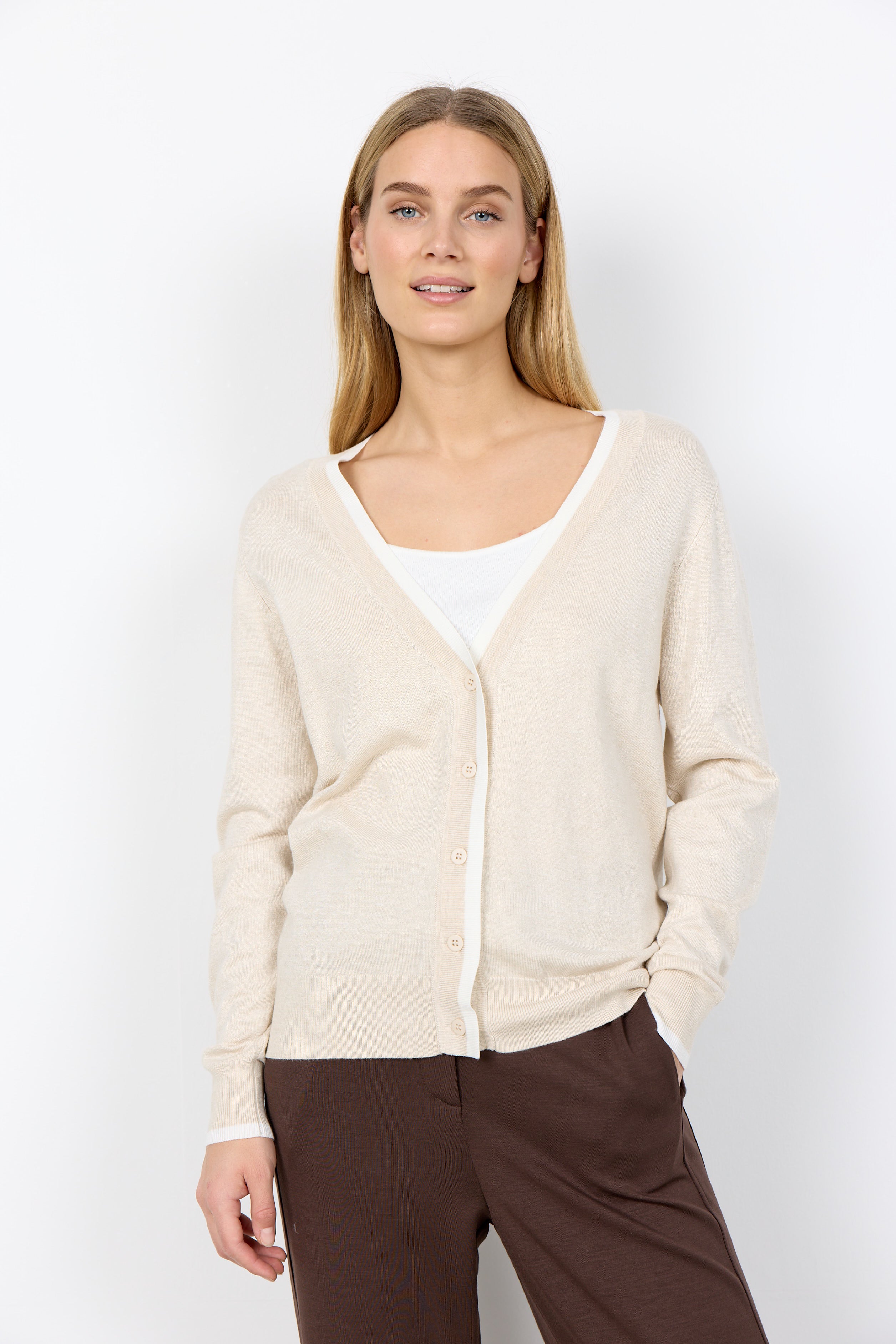 Woman wearing a beige cardigan over a white top with brown pants on a white background