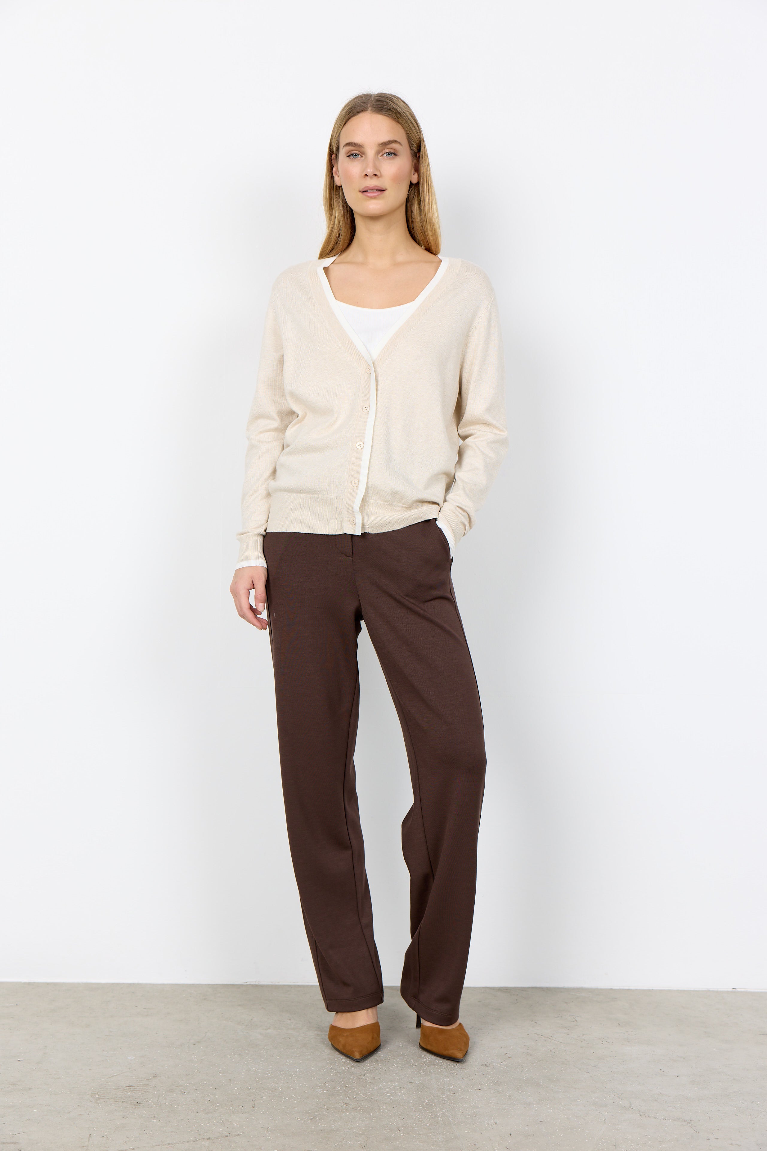 Woman wearing a beige cardigan and brown pants against a white background