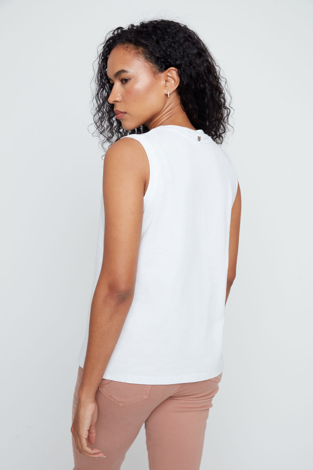 Woman wearing a white sleeveless top and brown pants against a plain background